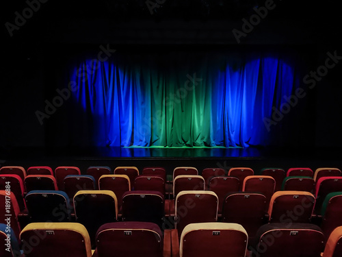 The auditorium in the theater. Blue-green curtain on the stage. Multicolored spectator chairs. Lighting equipment.