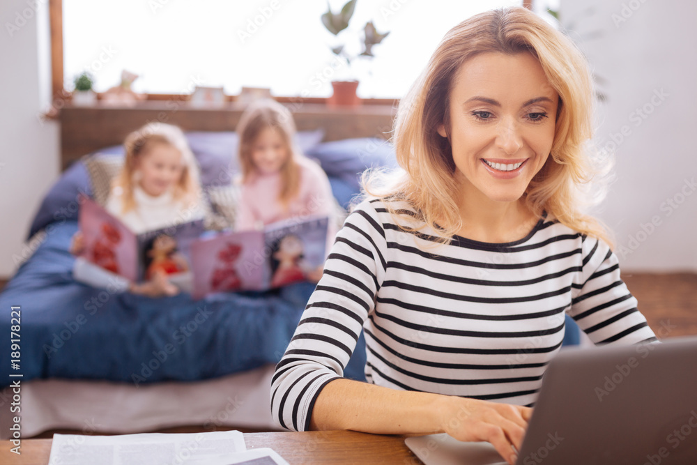 Working. Attractive content blond young mother smiling and working on her laptop while her daughters reading magazines Working. Attractive content blond young mother smiling and working on her laptop while her daughters reading magazines
