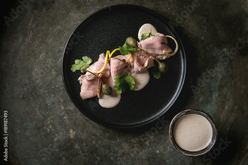 Vitello tonnato italian dish. Thin sliced veal with tuna sauce, capers and coriander served on black plate over old dark metal background. Top view, space photo