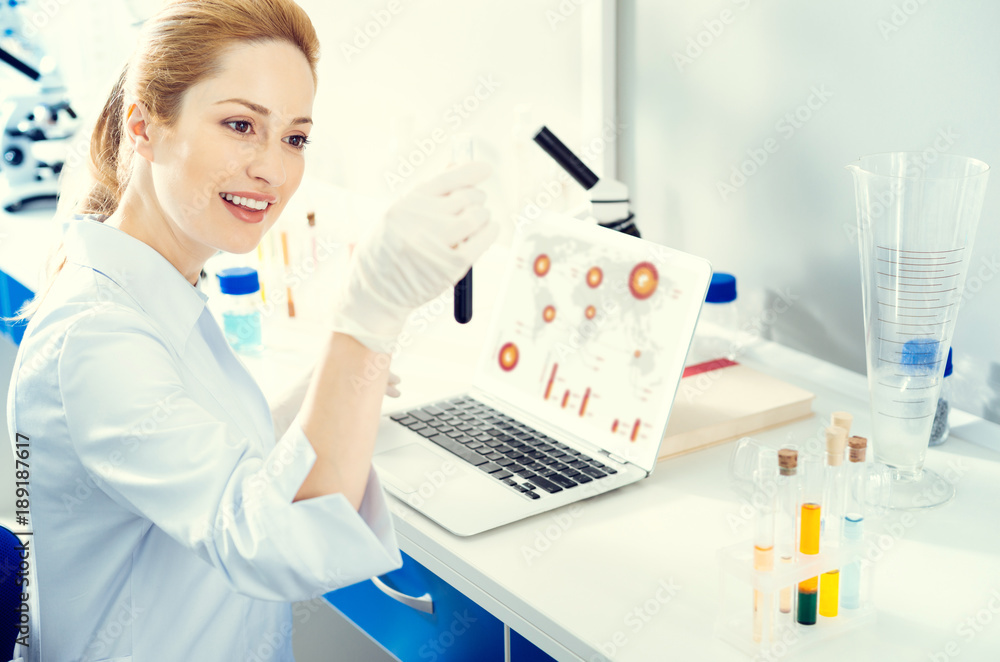 Up and coming professional. Selective focus on a female researcher sitting at a laptop and smiling while looking at a test tube in a lab. Up and coming professional. Selective focus on a female researcher sitting at a laptop and smiling while looking at a test tube in a lab.
