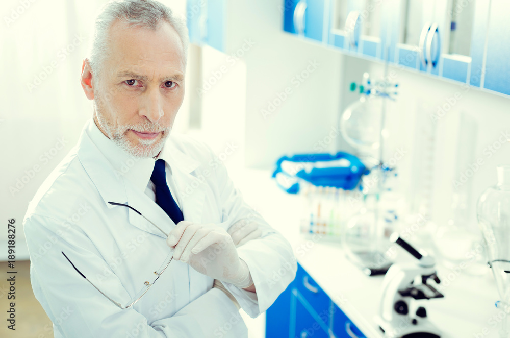 Experienced professional. Top view on a confident scientist holding his glasses and looking into the camera while standing in a laboratory. Experienced professional. Top view on a confident scientist holding his glasses and looking into the camera while standing in a laboratory.