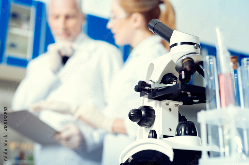 Professional equipment. Selective focus on a modern microscope standing on a lab table while mature colleagues discussing something in the background. Professional equipment. Selective focus on a modern microscope standing on a lab table while mature colleagues discussing something in the background.