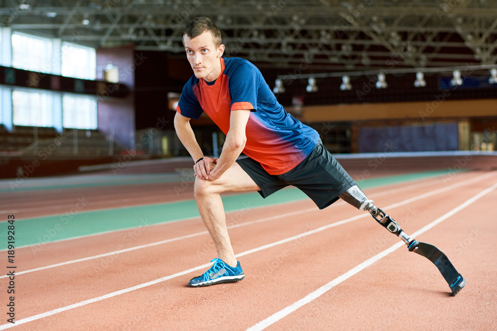 Full length portrait of young amputee sportsman  warming up before running practice in modern gym stretching legs on track, copy space