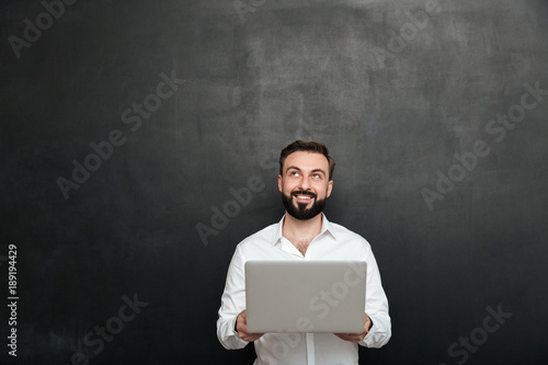 Portrait of smiling bearded man holding silver personal computer and looking upward, isolated over dark gray wall