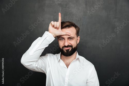 Portrait of unhappy guy 30s posing on camera and showing loser sign on his forehead, over graphite background photo