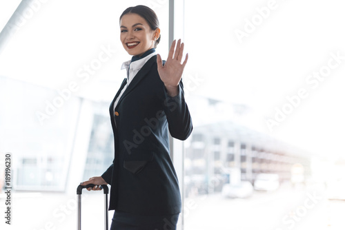 attractive young stewardess with suitcase waving at camera in airport photo