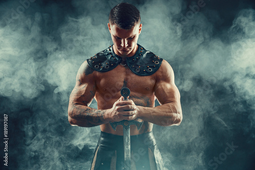 Portrait of handsome muscular gladiator with sword. Studio shot. Smoke on background.