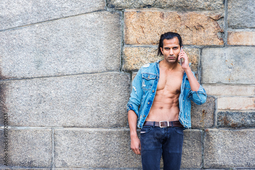 Εκτύπωση καμβά Young East Indian American Man wearing blue fashionable Denim jacket, opened, showing half naked body, black jeans, standing against rock wall in New York, seriously talking on cell phone
