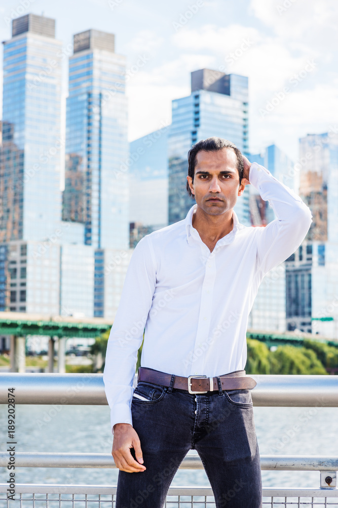 Young East Indian American Man wearing white shirt, black jeans, standing  in business district with high buildings by Hudson River in New York, hand  on back of head, looking forward.. Stock Photo