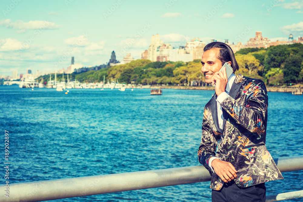 Young East Indian American Man wearing black fashionable patterned blazer, standing by Hudson River in New York, smiling, talking on cell phone. Harbor with boats on background..