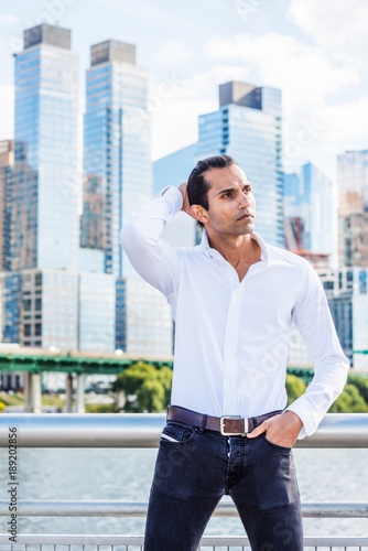Young East Indian American Man wearing white shirt, black jeans, standing in business district with high buildings by Hudson River in New York, hand on back of head, looking around, thinking..