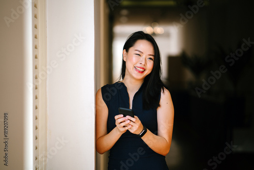 Free Download Portrait of a professional businesswoman standing in the middle of a corridor and looking up from her smartphone and smiling. - GFXLoad
