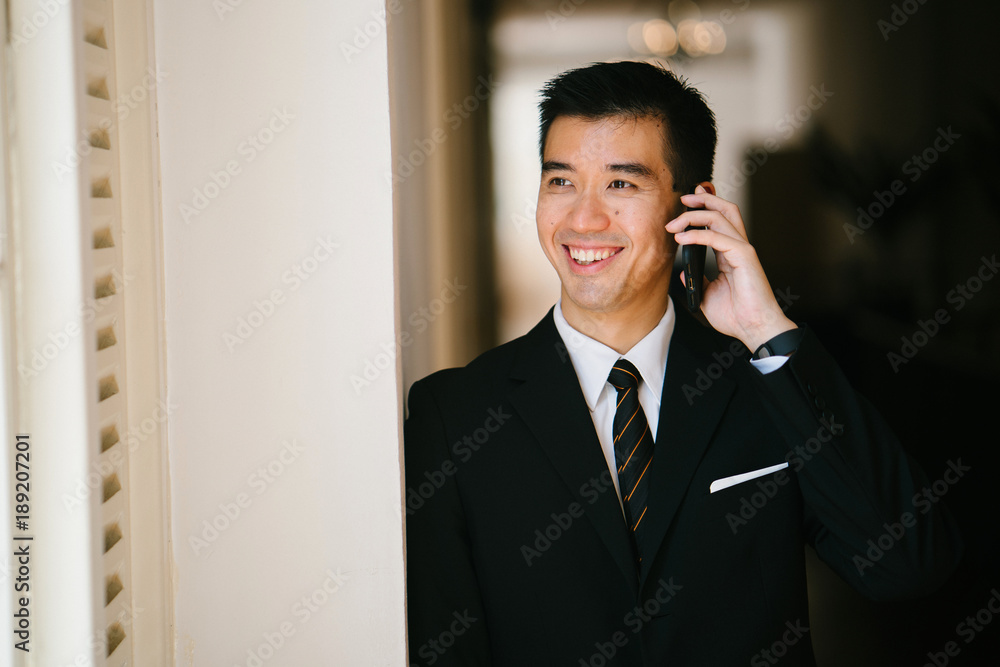 Portrait of young handsome man standing in a well lit corridor. He is wearing formal business attire and talking and smiling on his phone. Portrait of young handsome man standing in a well lit corridor. He is wearing formal business attire and talking and smiling on his phone.