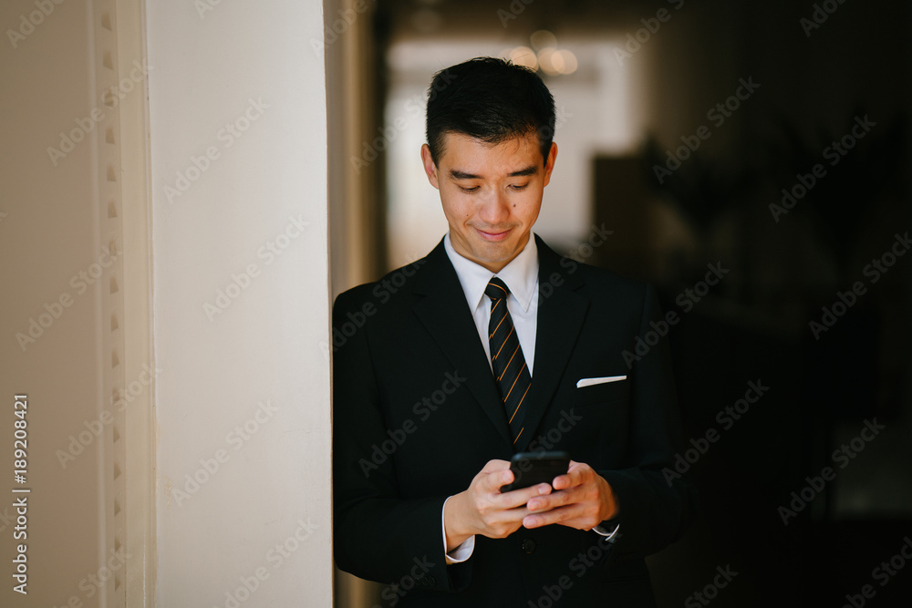 Portrait of young handsome man standing in a well lit corridor. He is wearing formal business attire, looking and smiling at his phone. Portrait of young handsome man standing in a well lit corridor. He is wearing formal business attire, looking and smiling at his phone.