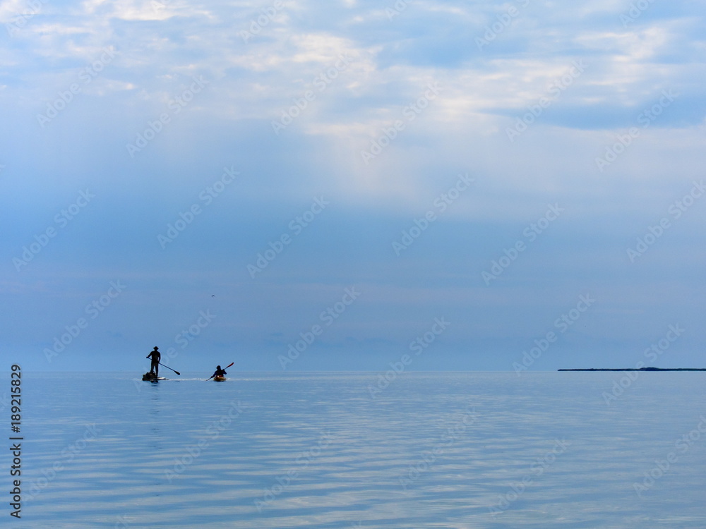 Silhouette of stand up paddle boarder and kayak paddling on a flat warm quiet sea. Two water tourists on stand up paddle boarder (SUP) and yellow kayak paddling on a flat warm quiet sea