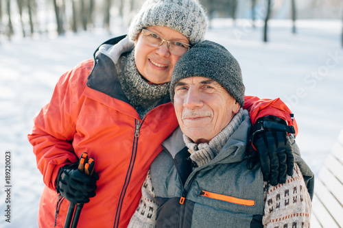 Obraz na płótnie Senior couple embracing in winter snowy park. Mature woman and smiling old man hugging looking at camera. w salonie