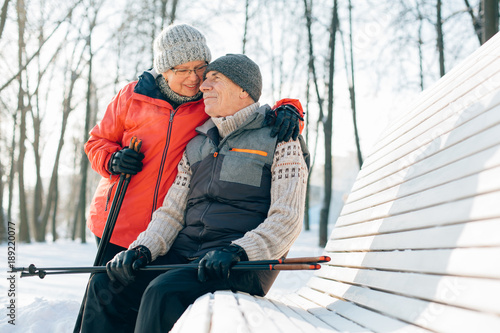 Pretty senior couple sitting with nordic walking poles in winter park. Mature woman and old man resting outdoors. Healthy lifestyle concept.