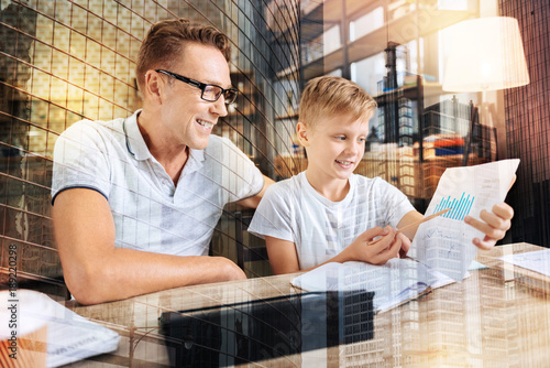 Lets compare. Cheerful kid expressing positivity while demonstrating his project and sitting near his father