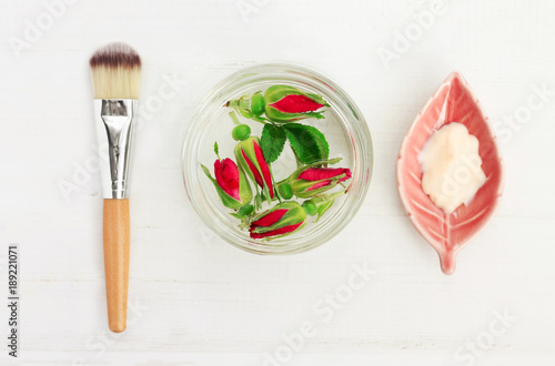 Glass jar of rose water with flowers, facial herbal mask, cosmetic application brush. Ingredients for home spa viewed from above white woode background.