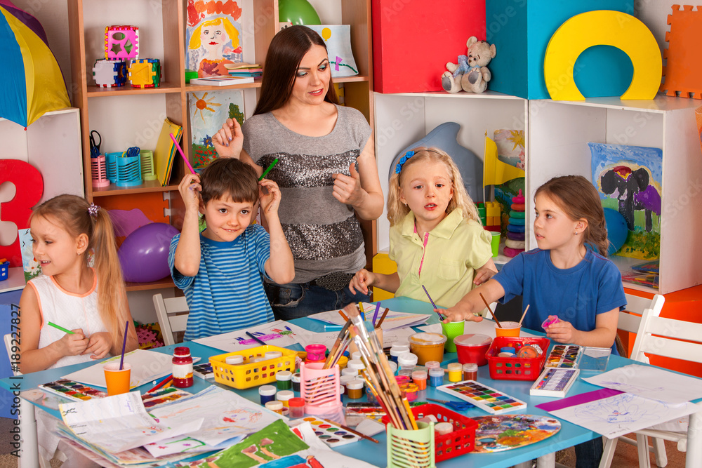 Fototapeta premium Kids playroom organization of children painting and drawing in kid's club. Craft lesson in primary school. Kindergarten teacher and students work together. Teacher is unhappy with children.
