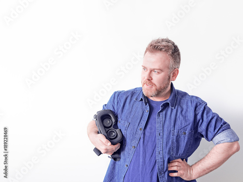 A cute red bearded guy in a blue denim shirt looks into the distance and holds in his hand a helmet of virtual or augmented reality. Image on the topic of modern technology, games or training