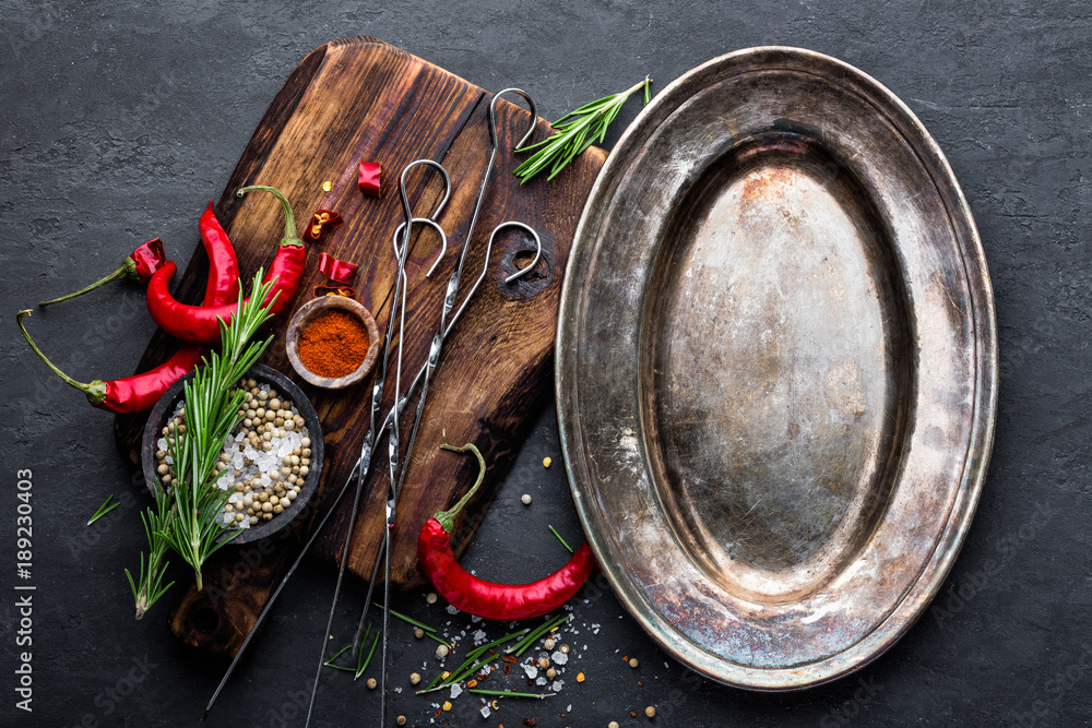 Culinary background with empty metal plate and space for a text. Flat lay composition of chili peppers, rosemary, spices and skewers for shish kebab on a black surface, top view