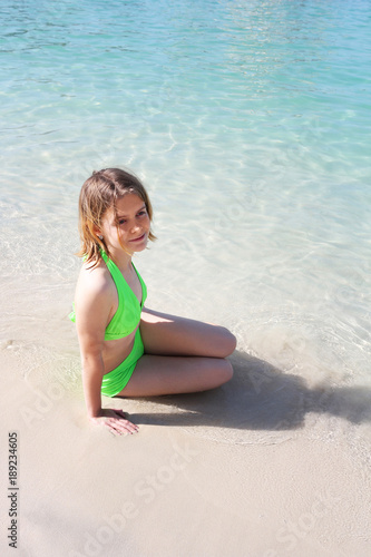 Young girl in a lime-colored swimsuit sitting on the beach by the water. Clear calm sea. White sand. Sunny day. Good mood