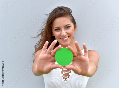 A young girl stands in front of a grey cement wall. She holds up a round green card to the camera. She shows a positive expression on her face. Green for go is the message she is sending.