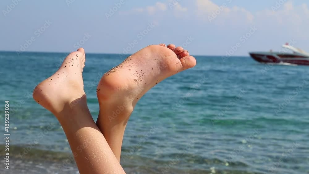 LIttle child legs isolated at blue sea water and blurry sailing motorboat in background. Closeup of small barefoot kid relaxing down with his belly at summer beach. Holidays and vacation concept. LIttle child legs isolated at blue sea water and blurry sailing motorboat in background. Closeup of small barefoot kid relaxing down with his belly at summer beach. Holidays and vacation concept.