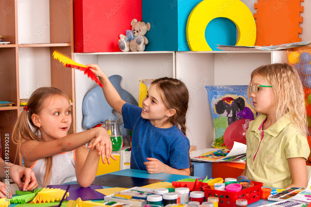 Fototapeta premium School children with scissors in kids hands cutting paper with teacher in class room. Children's project in kindergarten zone. Large group girls and boys together. Team of students makes paper fans.