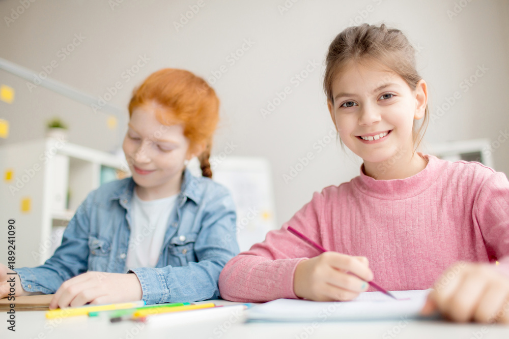 Cute girl with toothy smile looking at camera at lesson with her classmate near by Cute girl with toothy smile looking at camera at lesson with her classmate near by