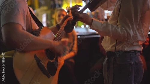 young people are celebrating in the pub. two men play the guitar and violin. live music in the bar. people are sitting on high taksoyah against the back of the bar. Holiday of drink and beer. cheerful