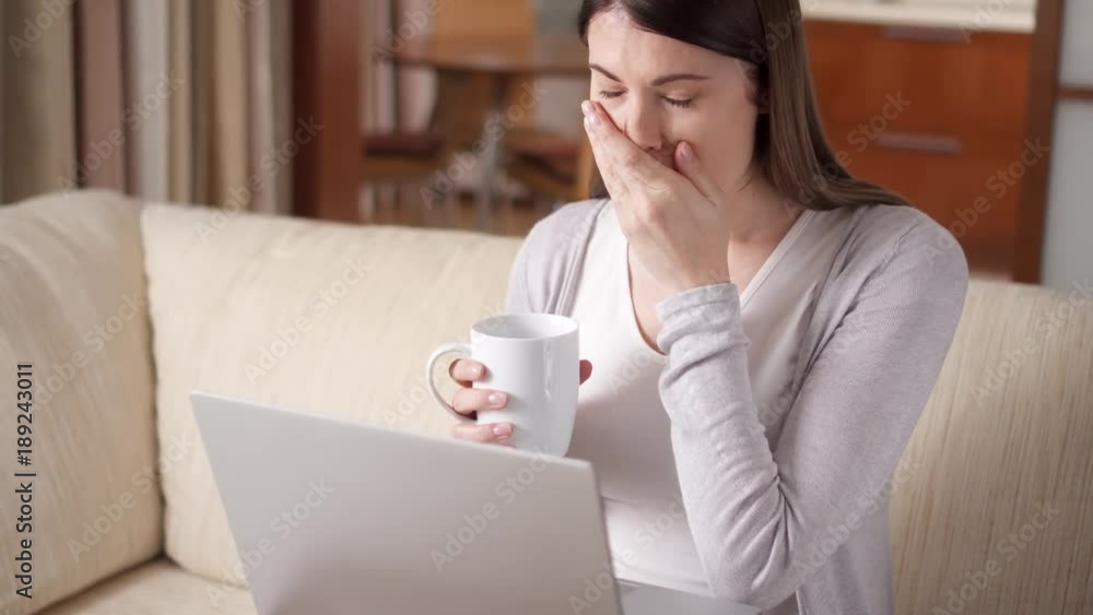 Sleepy tired yawning businesswoman working on laptop computer at home drinking coffee from cup. Young casual female professional working on computer device in the morning. Home-office concept. Sleepy tired yawning businesswoman working on laptop computer at home drinking coffee from cup. Young casual female professional working on computer device in the morning. Home-office concept.
