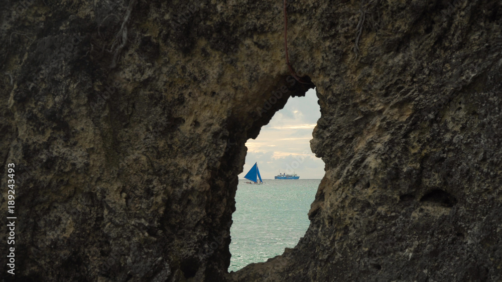 Sailing boat view through a hole in the rock. Sailing ship yachts with blue sails in the ocean. Sail boat on sea. Philippines, Boracay. Travel concept.