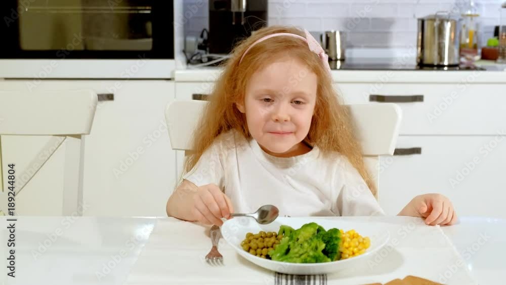A small beautiful girl eats broccoli and green peas with pleasure, at the table at home A small beautiful girl eats broccoli and green peas with pleasure, at the table at home