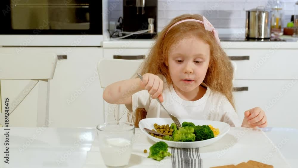 A small beautiful girl eats broccoli and green peas with pleasure, at the table at home A small beautiful girl eats broccoli and green peas with pleasure, at the table at home