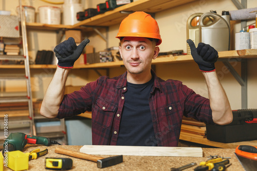 Handsome smiling caucasian young man in plaid shirt, black T-shirt, orange protective helmet, gloves working in carpentry workshop at wooden table place with piece of wood, hammer, different tools.