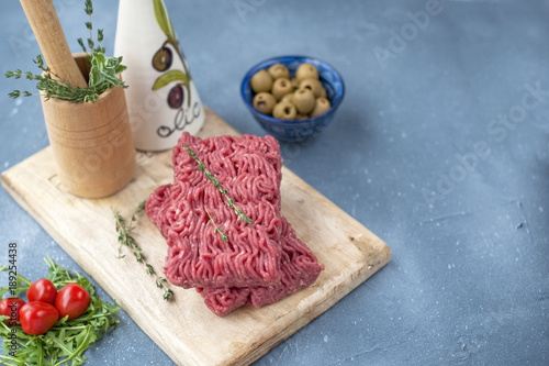 Fresh beef meat in forcemeat, on a wooden board. Raw eggs and tomatoes are small. Herbs for cooking. On a gray stone background. Free space for writing text.
