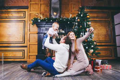 Christmas theme. Father throws the son of blond one year old, sit on a wooden tree in front of Christmas tree with gifts, mom makes selfie, self-portrait on the front camera phone of a silver color. photo