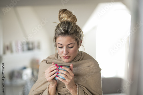 Beautiful middle-aged woman drinks tea from a glass in a cold living room dressed in a cotton becouse of cold winter. A sick girl drinks from a blue cup in the living room.