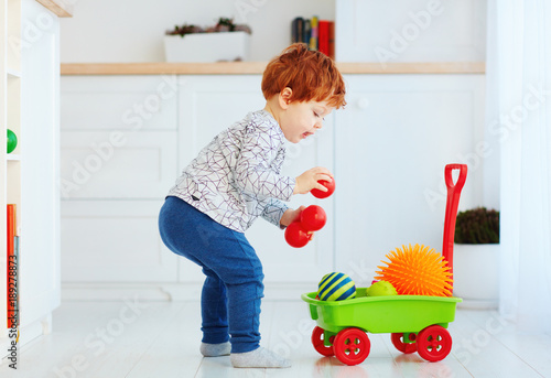 cute redhead toddler baby collecting different balls into toy pushcart photo