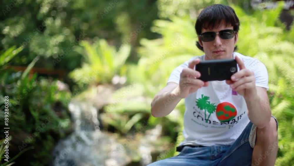 Man traveler wearing sunglasses with mobile phone sits near a waterfall in forest uses smartphone. 1920x1080, hd Man traveler wearing sunglasses with mobile phone sits near a waterfall in forest uses smartphone. 1920x1080, hd