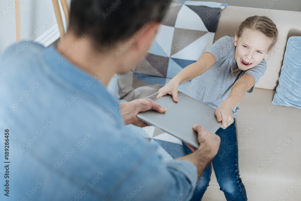 Obraz premium Give me. Angry fair-haired little girl shouting and taking away her laptop while daddy holding it