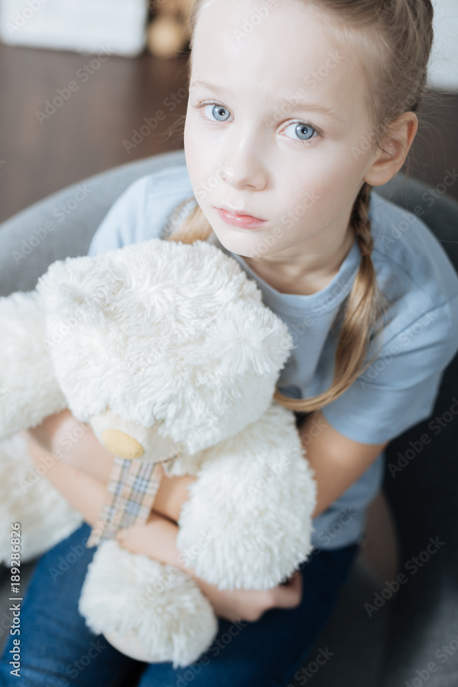 Loneliness. Unsmiling fair-haired little girl hugging and holding her teddy bear while sitting in the armchair Loneliness. Unsmiling fair-haired little girl hugging and holding her teddy bear while sitting in the armchair