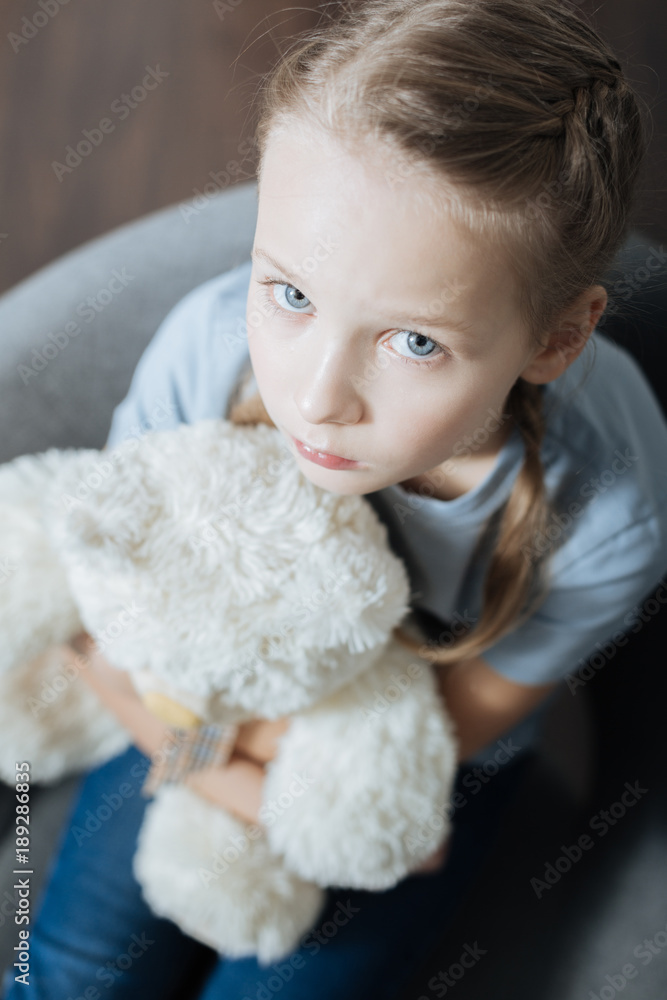 Being alone. Unsmiling blue-eyed little girl hugging and holding her teddy bear while sitting in the armchair Being alone. Unsmiling blue-eyed little girl hugging and holding her teddy bear while sitting in the armchair