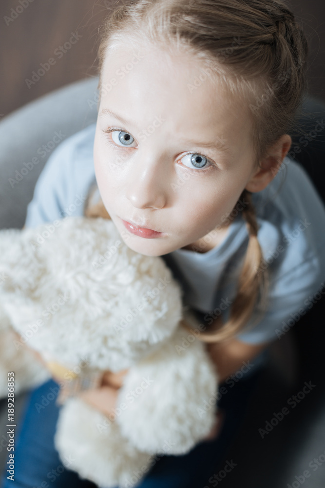 Feeling alone. Unhappy blue-eyed little girl hugging and holding her teddy bear while sitting in the armchair Feeling alone. Unhappy blue-eyed little girl hugging and holding her teddy bear while sitting in the armchair