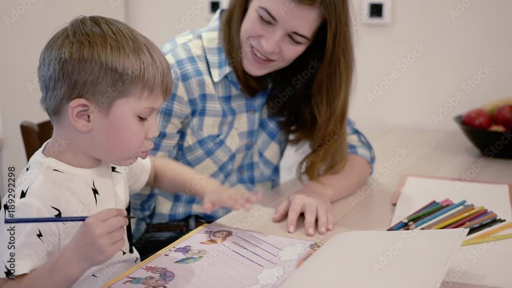 Side View - Beautiful Young Mother In Blue Shirt Helps Her Little Cute Son To Do Developmental Activities In A Notebook For The Preparation Of Preschoolers At Home. Slider Down to Up Shot Side View - Beautiful Young Mother In Blue Shirt Helps Her Little Cute Son To Do Developmental Activities In A Notebook For The Preparation Of Preschoolers At Home. Slider Down to Up Shot