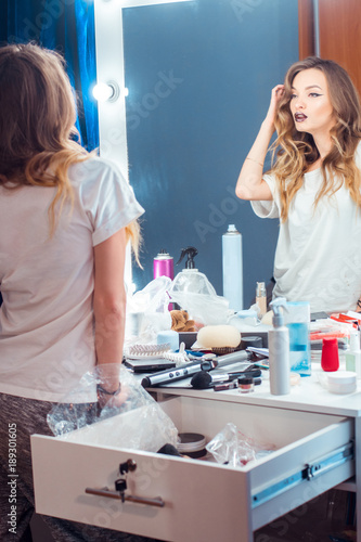 Young gorgeous woman with curly hairstyle and beautiful makeup looking at the mirror in hair salon