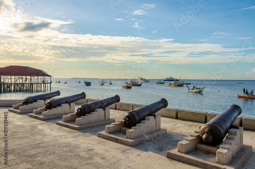 Five cannons in line at the waterfront in Stone Town Zanzibar. Several fishing boats are anchored off shore in the ocean. It's a tranquil scene at sunset with few clouds in the sky