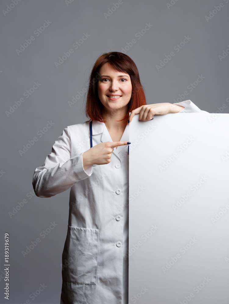 Image of smiling female doctor in white robe and with phonendoscope points finger at blank paper Image of smiling female doctor in white robe and with phonendoscope points finger at blank paper